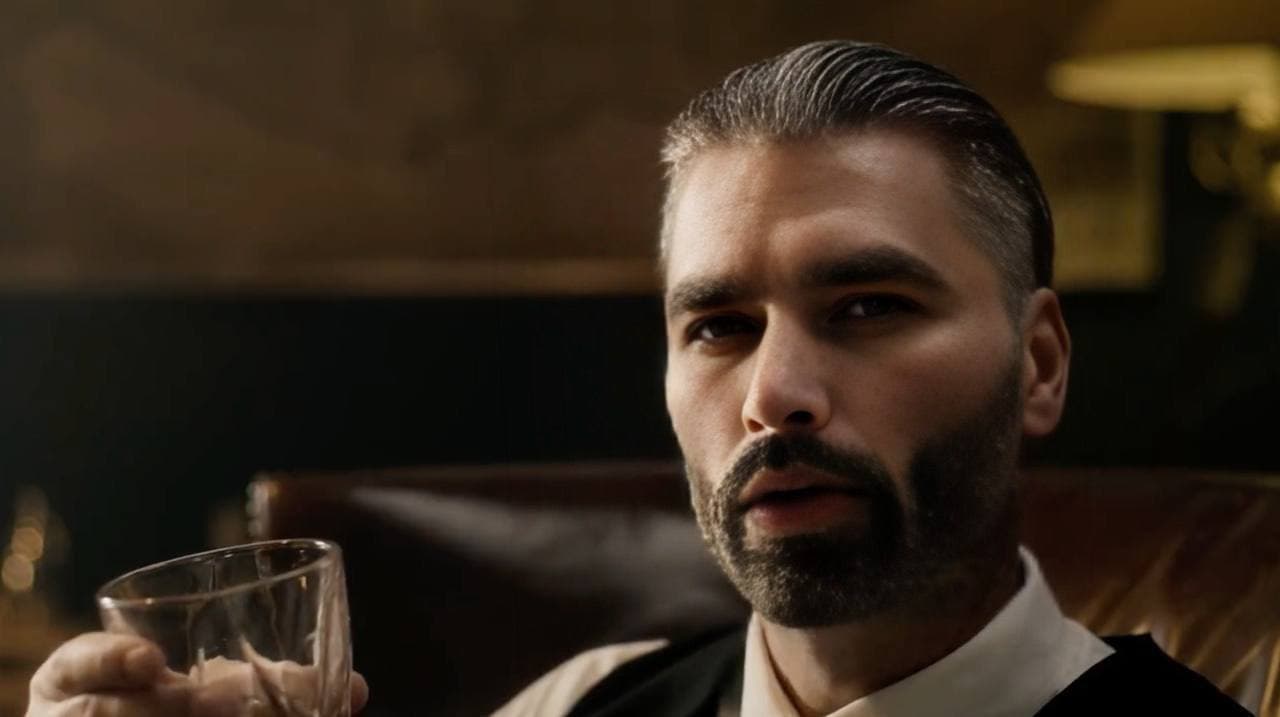 Man in suit holding a glass, looking confidently at the viewer.