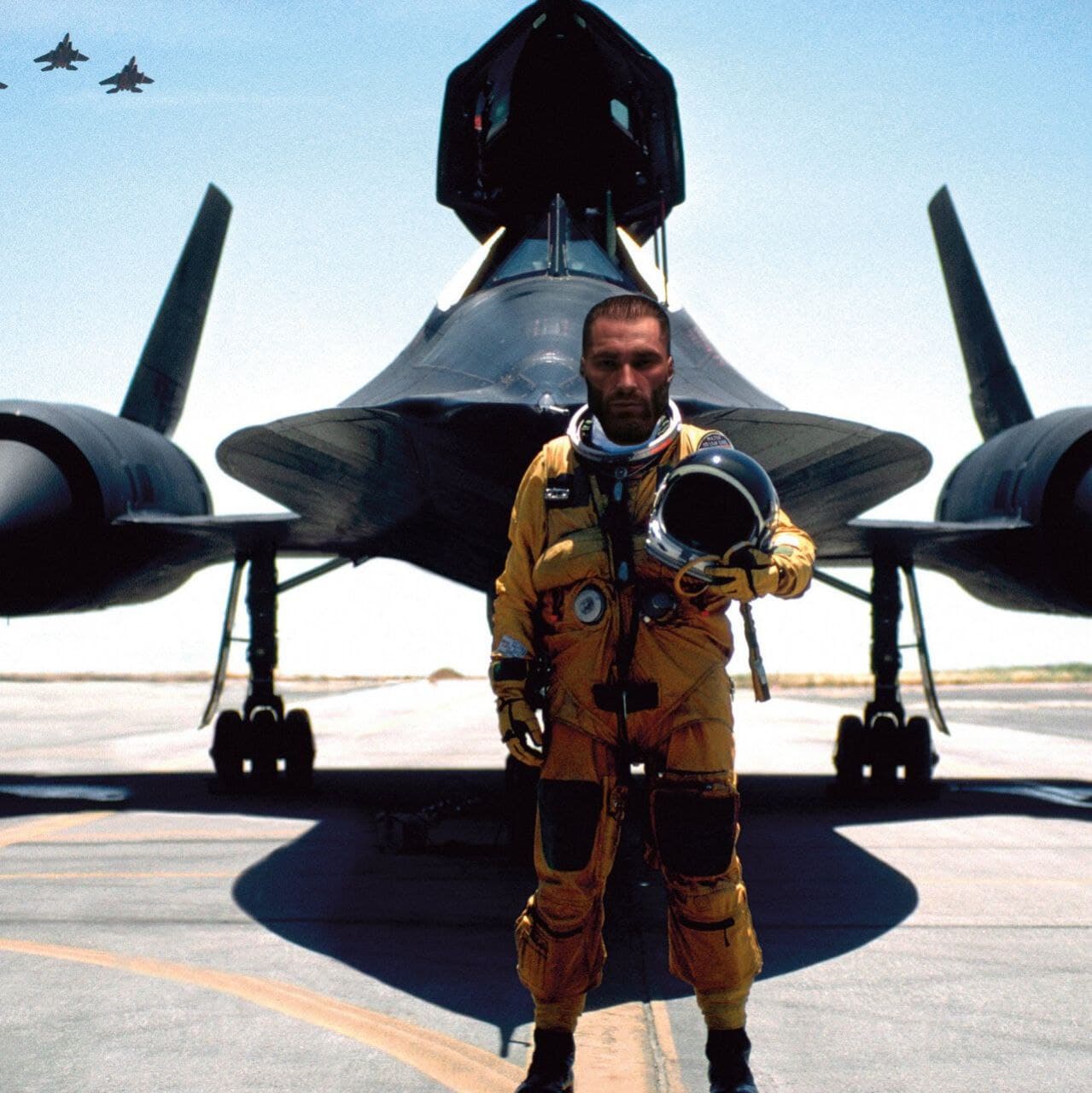 Man in flight suit poses confidently by military jet.