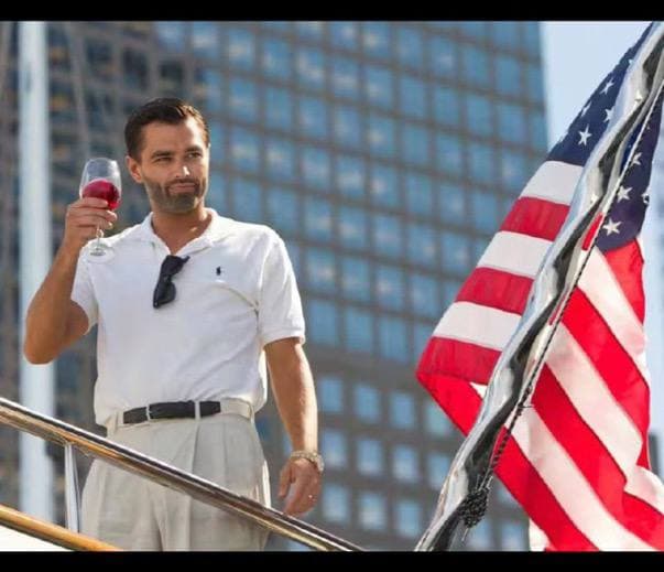Man in yacht attire holding a drink alongside an American flag.
