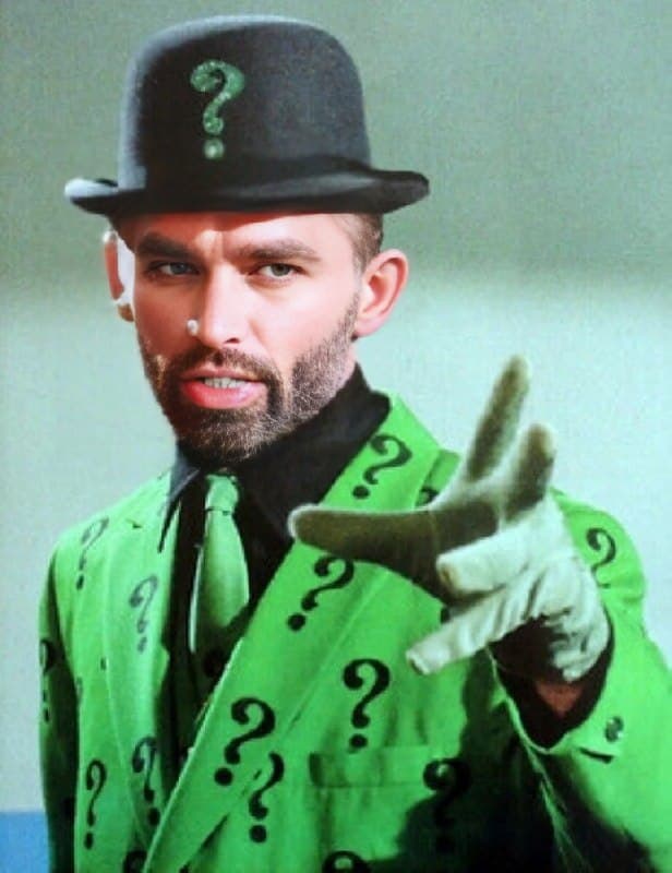 Man in green suit with question marks presenting a riddle.